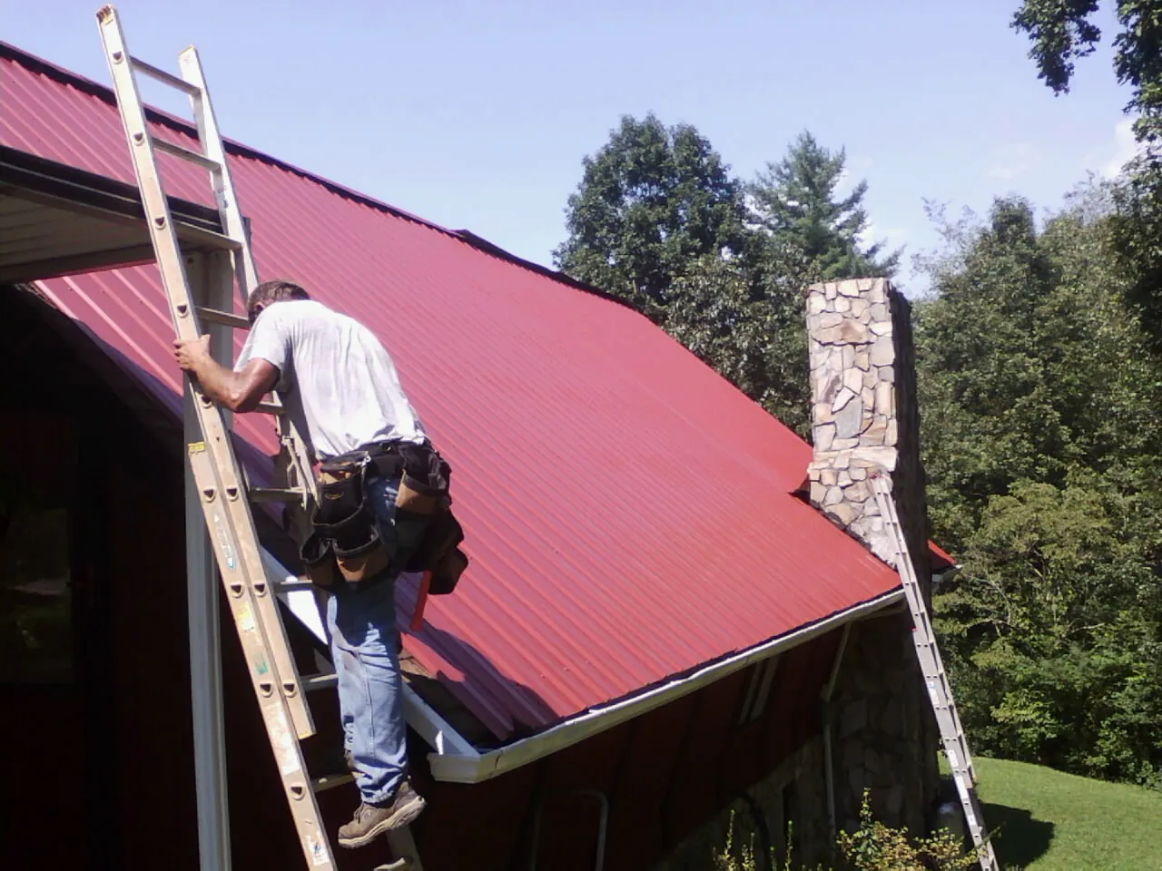 Roofer on a ladder beside a red metal roof and stone chimney — Soffit & Fascia Repair by Roofing Brothers in Porterdale